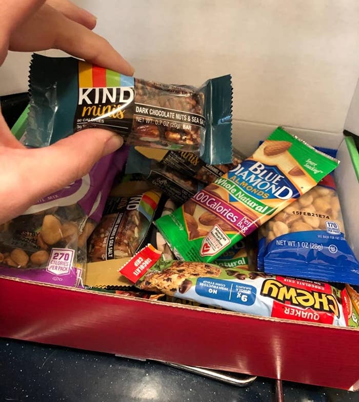 Reviewer is holding a Kind bar above a box of various snacks