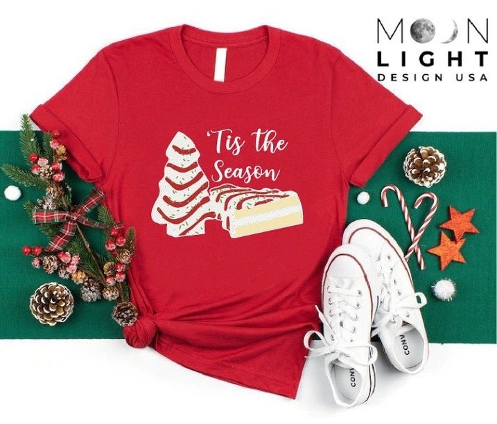 The red Little Debbie Christmas tree cake tee that says &#x27;Tis The Season, surrounded by holiday accessories
