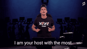 A GIF of a fitness instructor standing in front of an exercise bike slapping his hands to his chest as he says &quot;I am your host with the most&quot; in a Peloton video