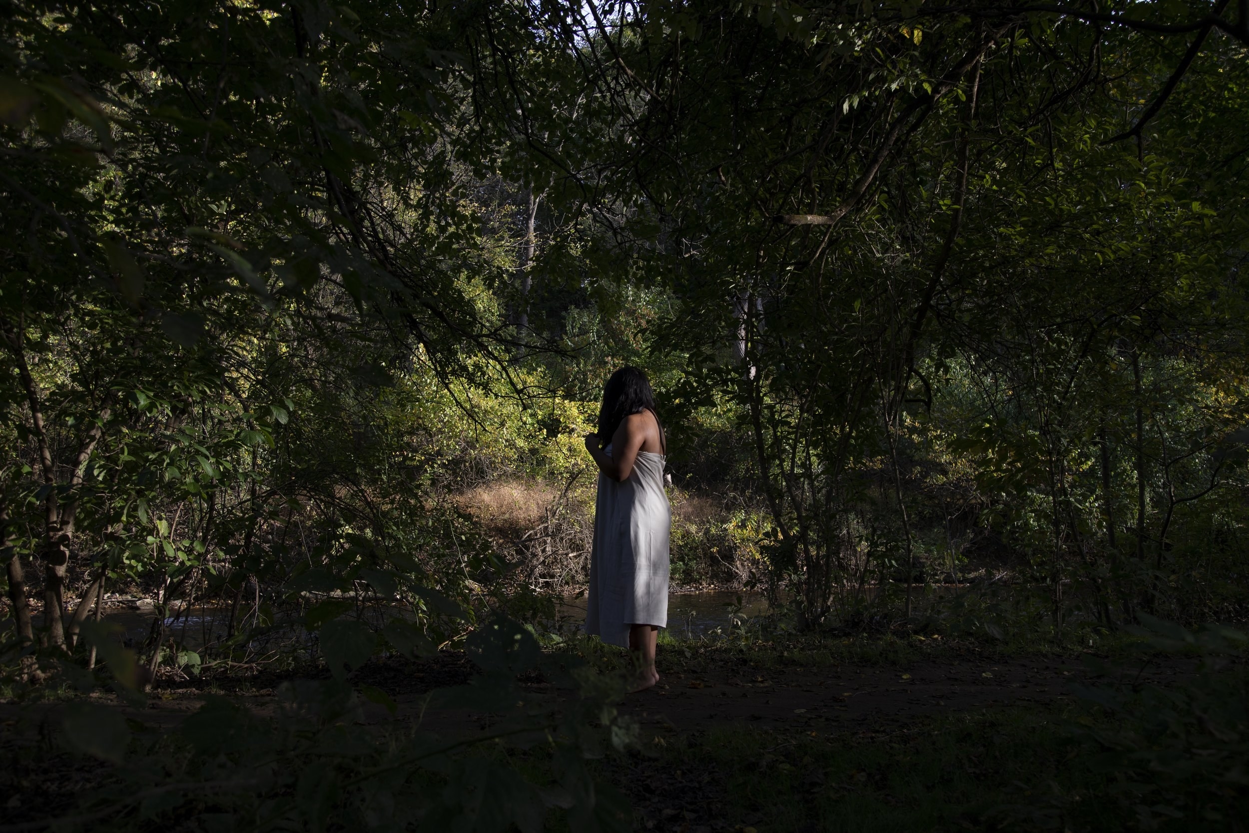 A person wrapped in a white sheet stands by a shaded creek