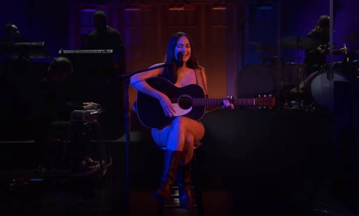 Kacey is naked while while sitting on a stool being covered up by a guitar