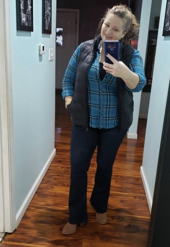 A reviewer&#x27;s selfie wearing the dark wash jeans with a flannel, vest, and boots