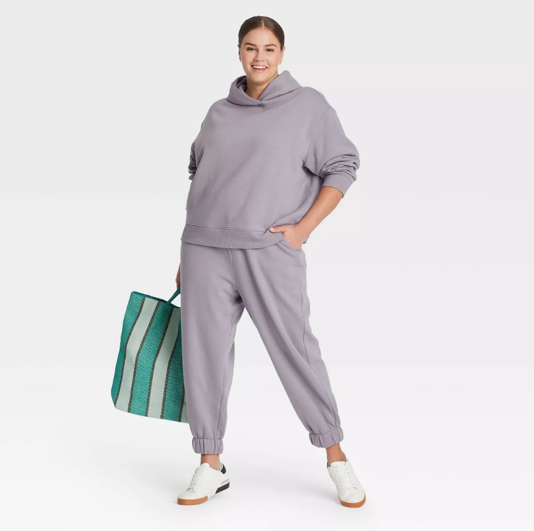 Model wearing gray hoodie with matching sweatpants, holding green striped bag