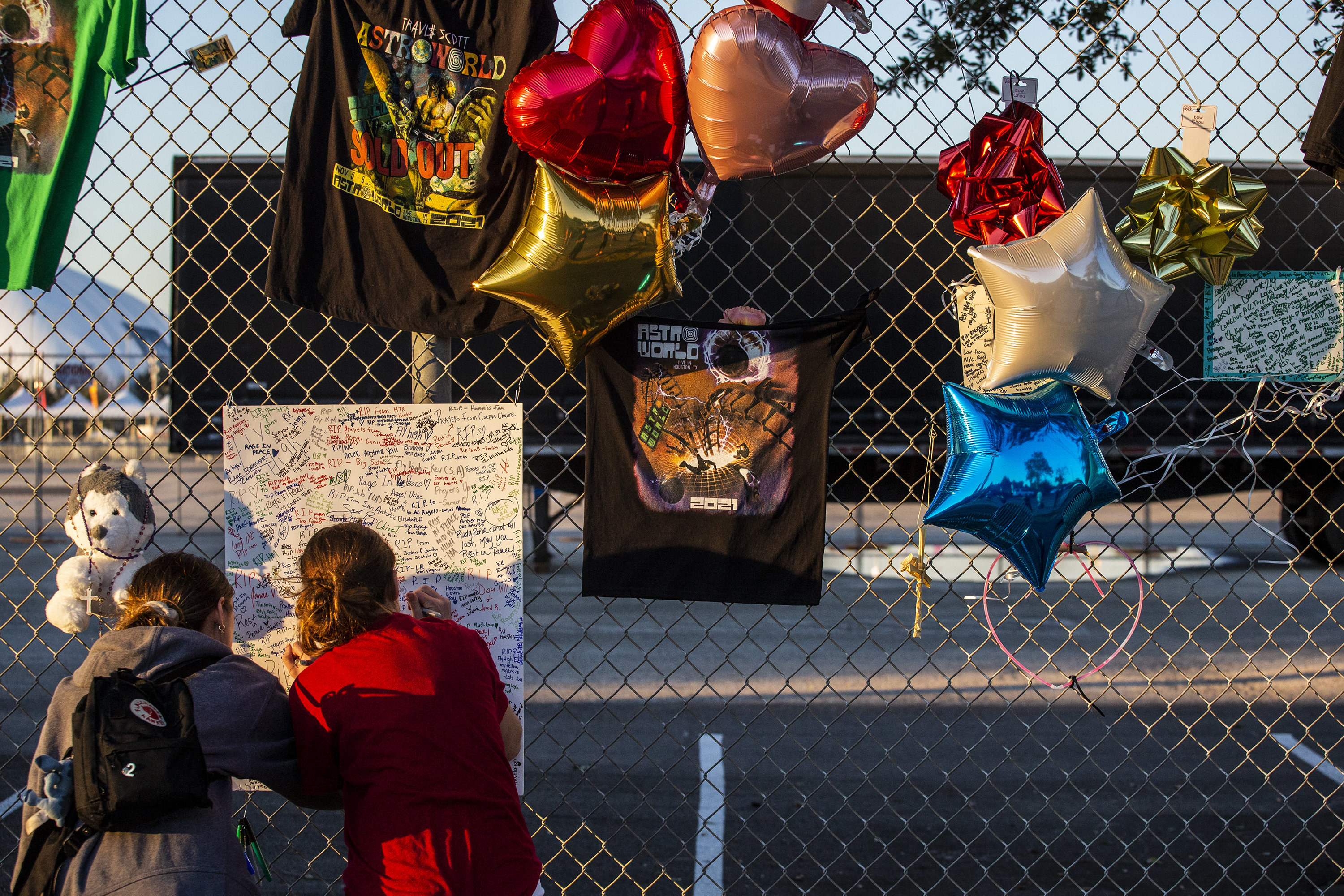 Two people write on a poster on a gate with balloons and other memorial items