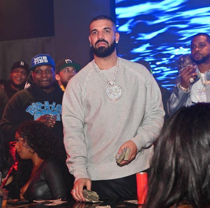 Drake holding wads of cash as he stands in front of a table surrounded by other people