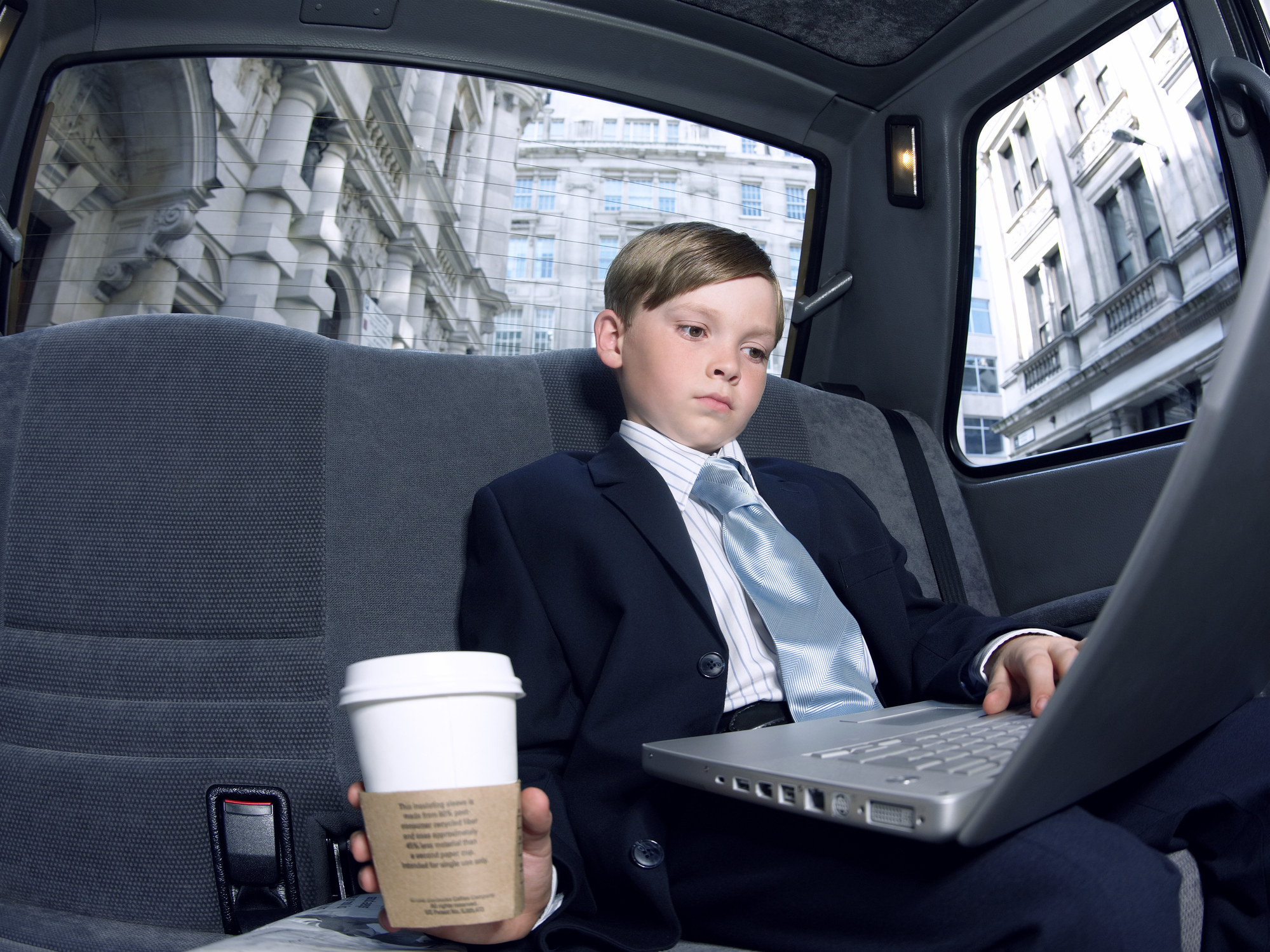 Boy dressed as a businessman, sitting in a taxi using a laptop computer