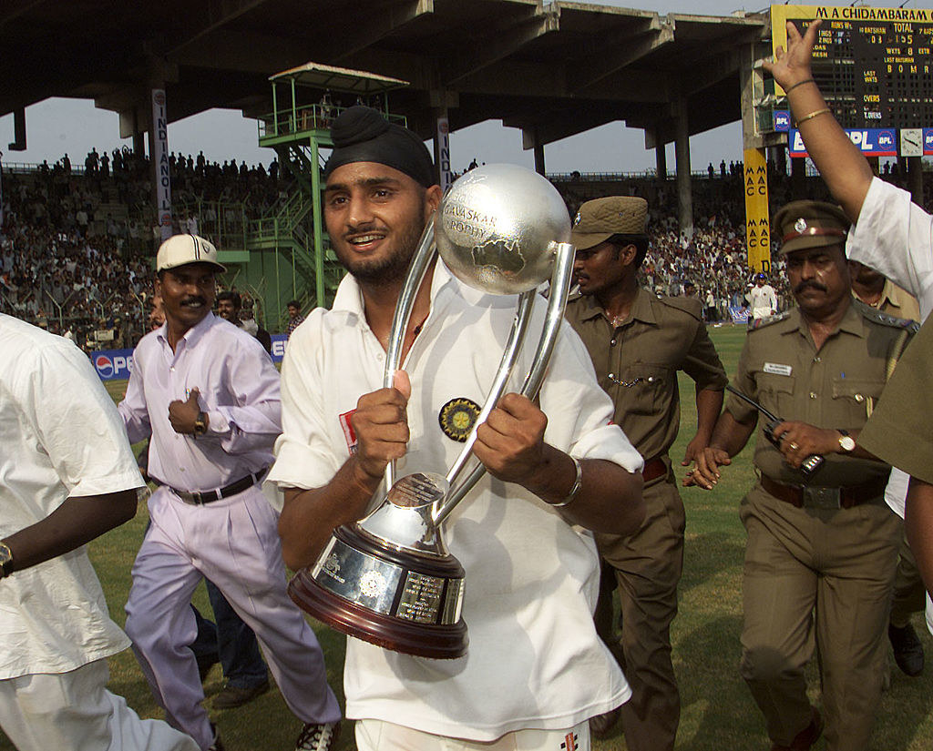 Harbhajan Singh lifts a massive trophy while smiling and walks on the cricket ground, surrounded by security personnel