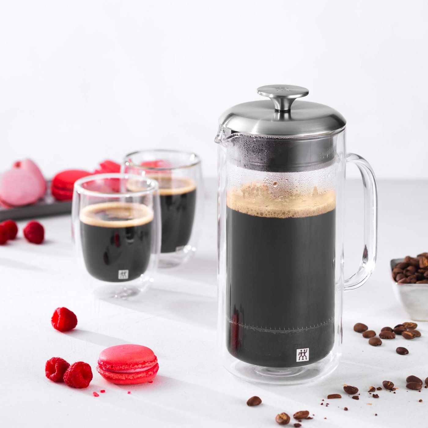 A filled French press sits on a table near raspberries, macarons and two cups of coffee
