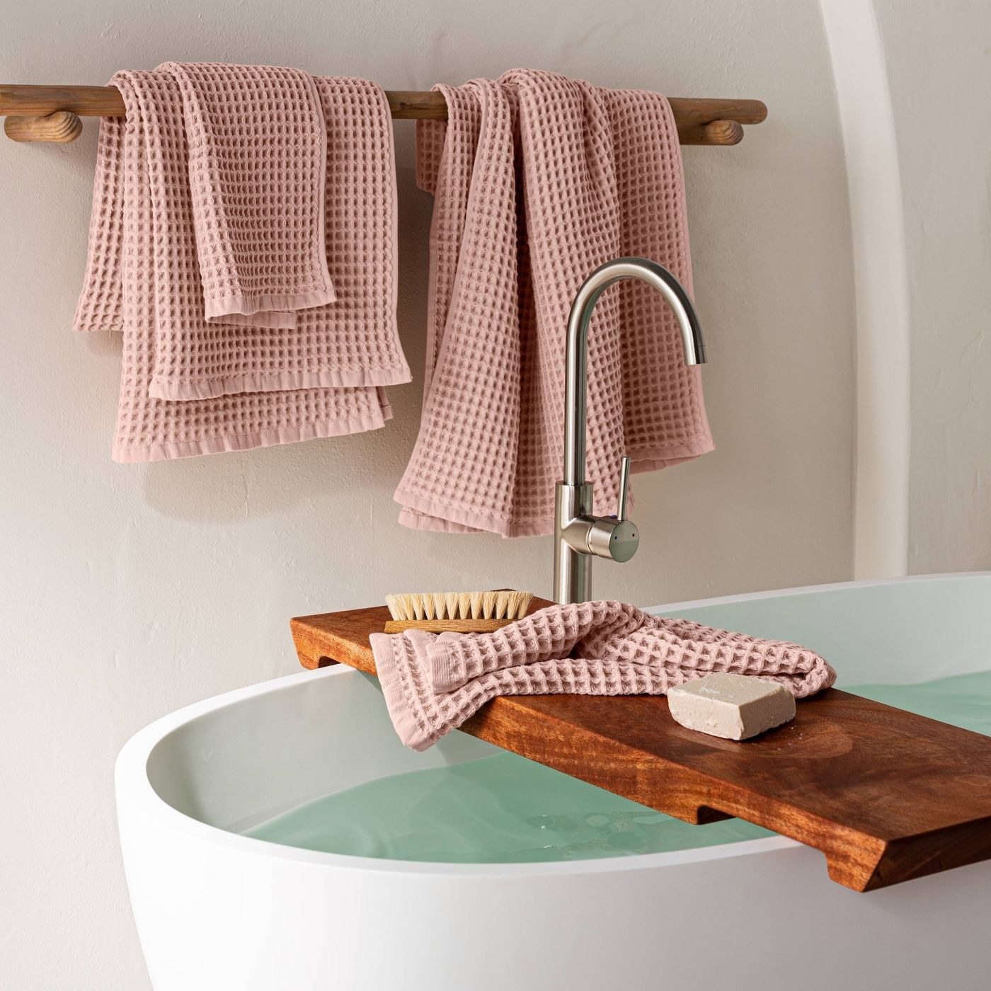 A deep bath filled with water with pink waffle towels draped over a wooden caddy