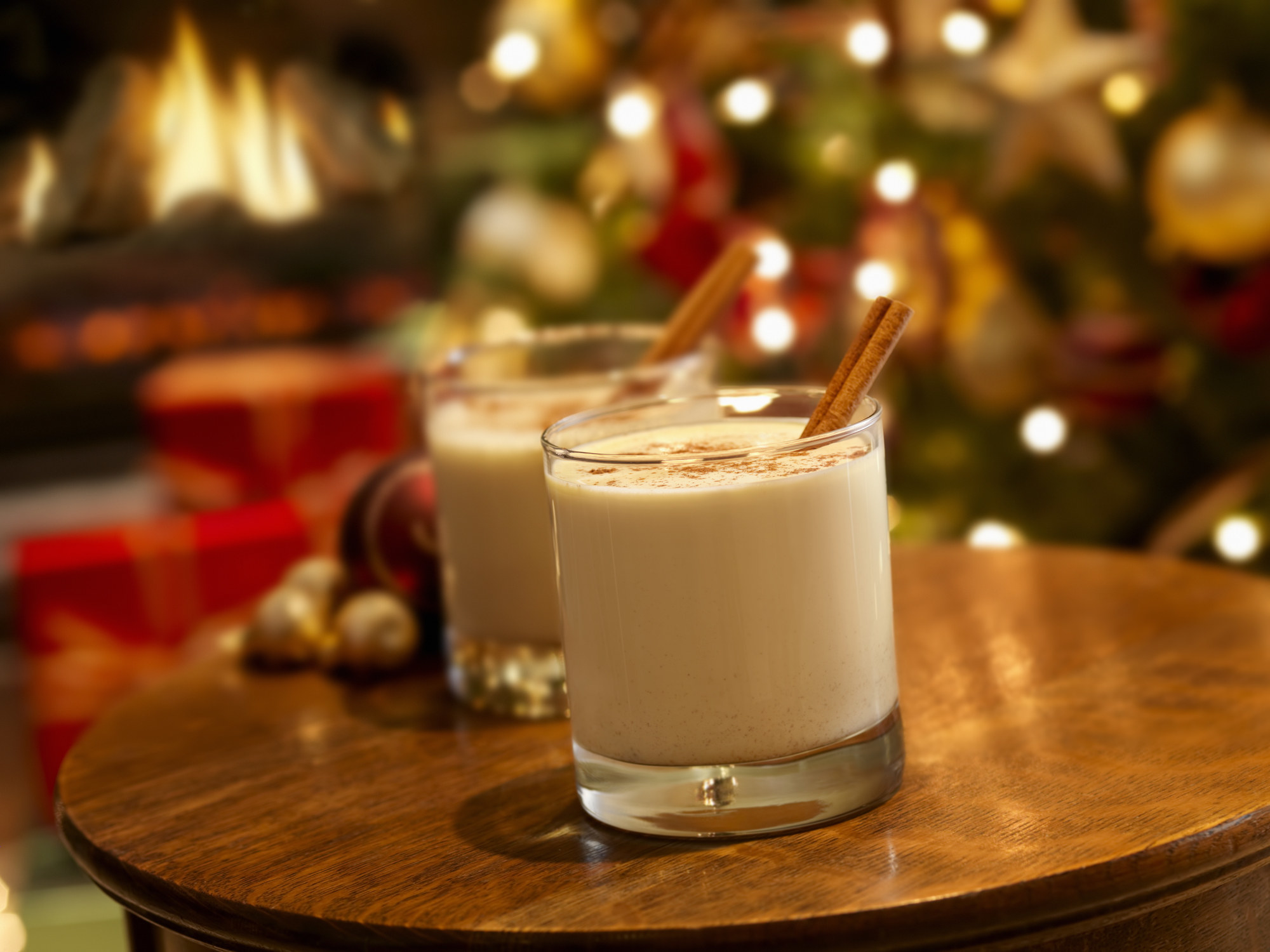 Two glasses of eggnog and cinnamon sticks on a table. There is a Christmas tree with lights on in the background.