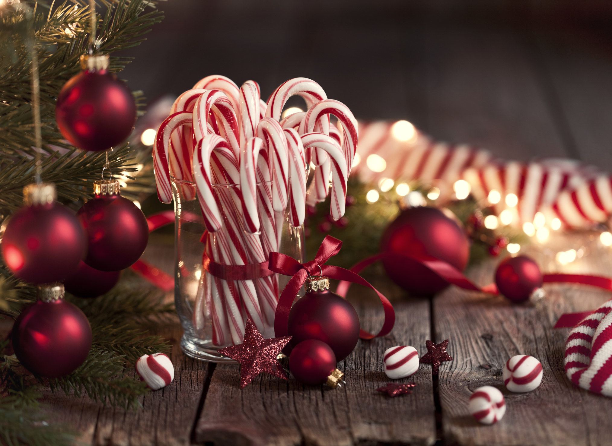 On a wooden table there is a glass with candy canes in it. Next to it is a christmas tree and there are red baubles everywhere.