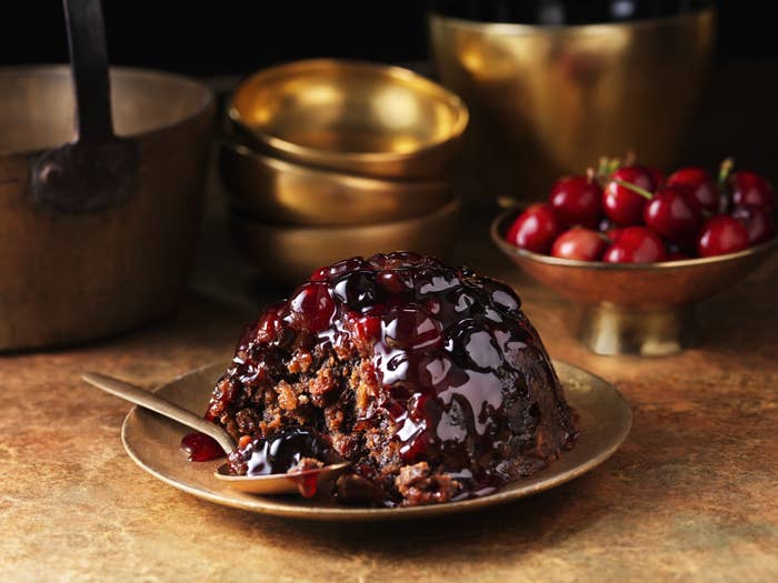 On a gold plate there is a Christmas pudding and a spoon. There are gold bowls in the background and a bowl of cherries.