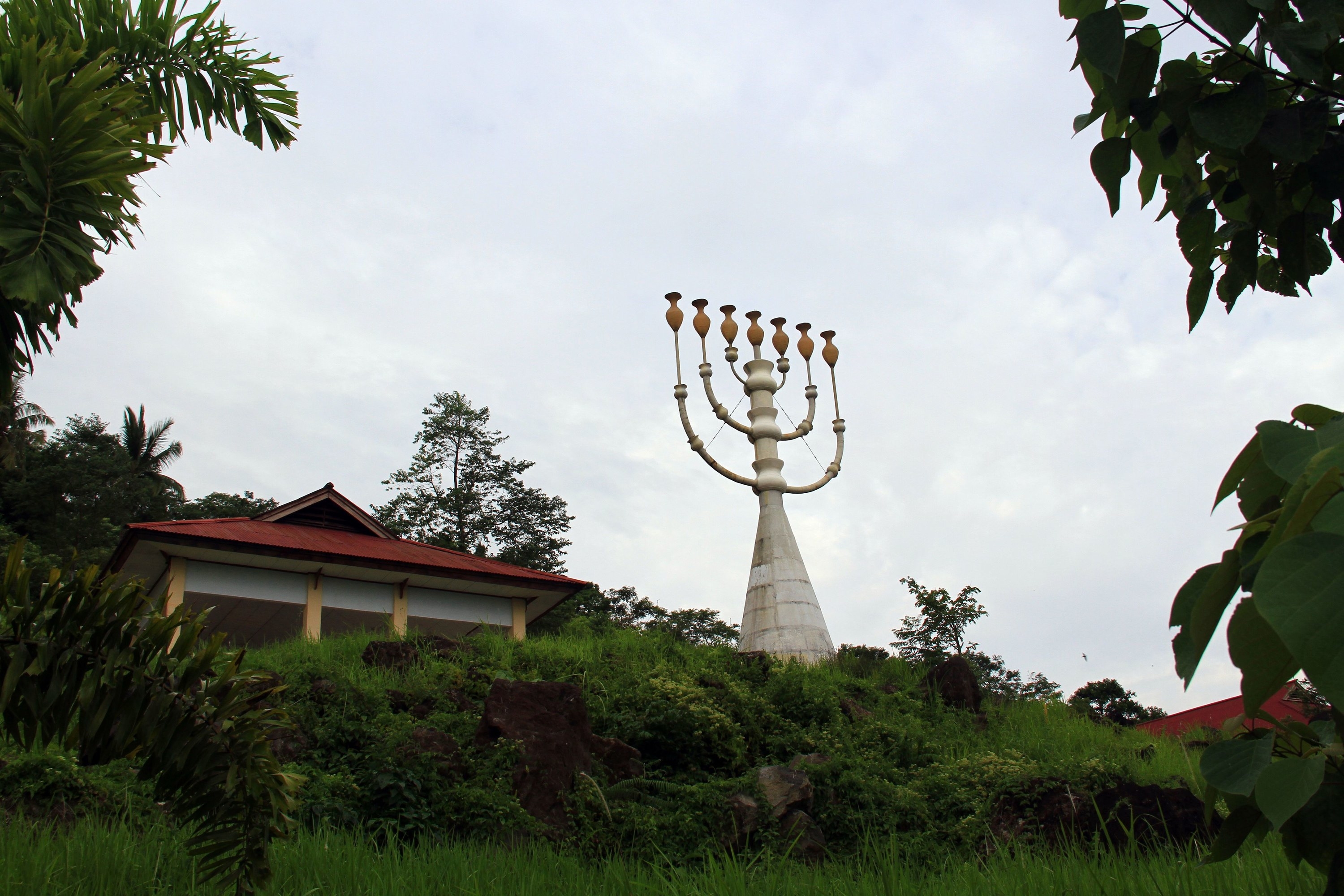 A large unlit menorah is photographed from below, next to a temple and with a lot of greenery 