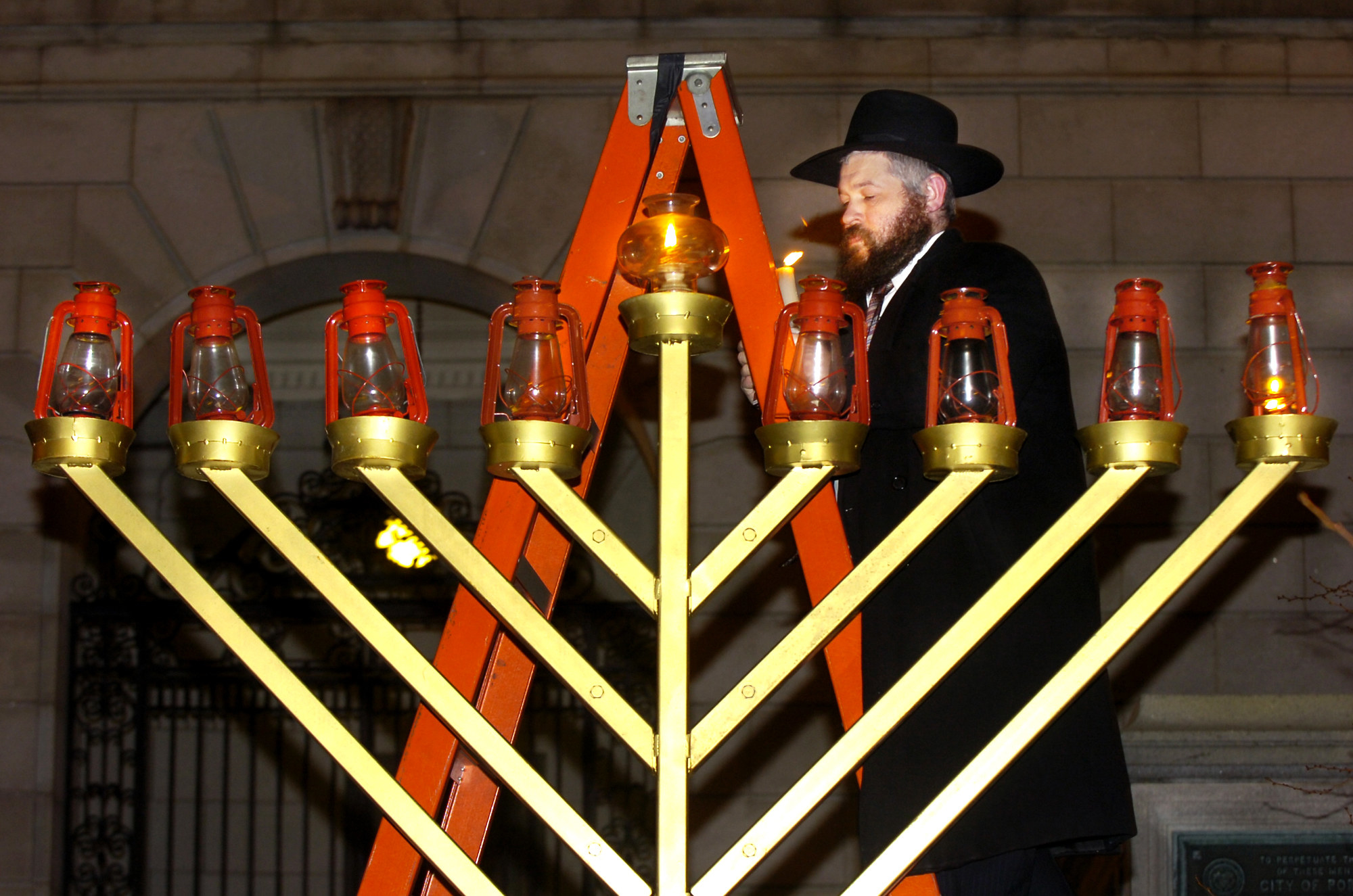 A man on an orange ladder lights one of eight lanterns on a giant menorah