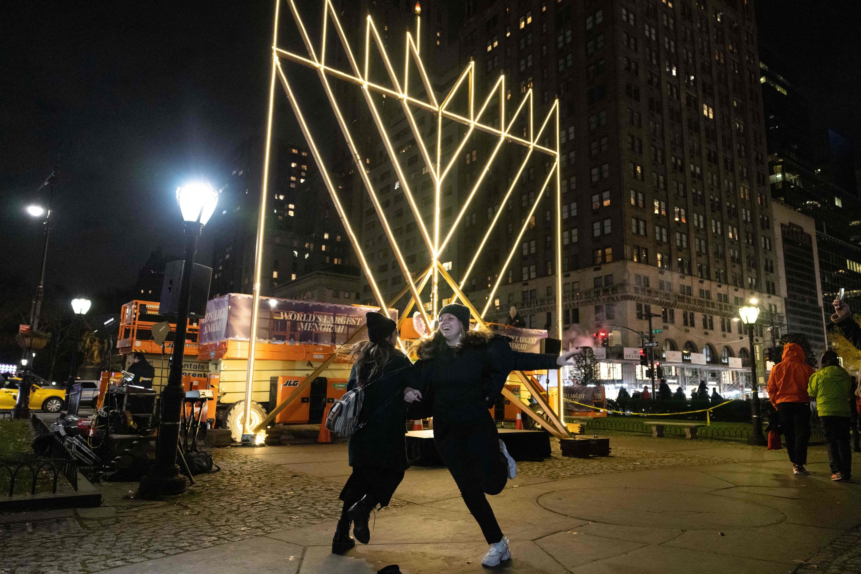 Two women dance outside a large menorah in brooklyn at night