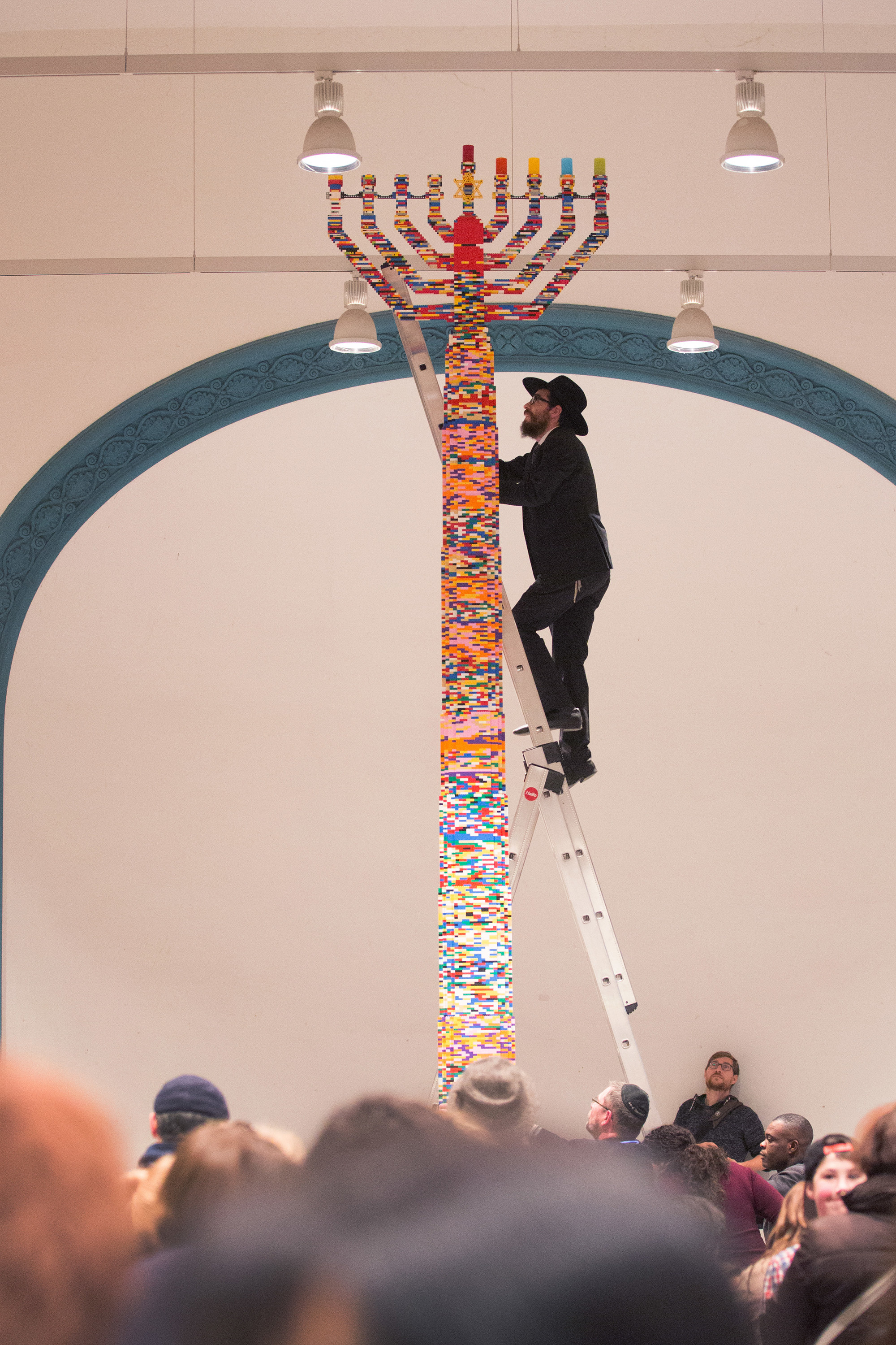 A rabbi climbs a ladder to get to the top of an extremely tall lego menorah in front of a crowd