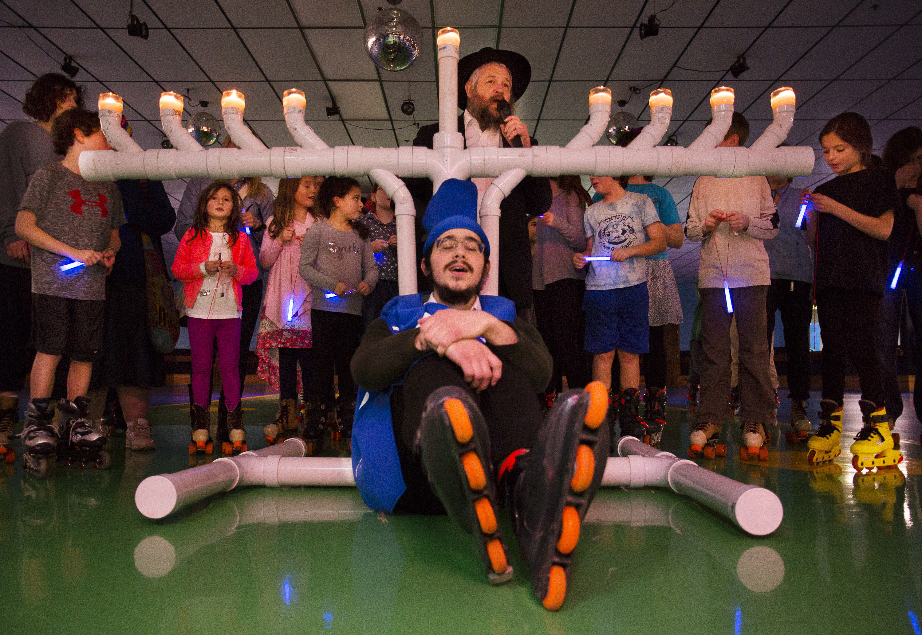 A man in roller skates site below a menorah made of PVC pipe, with a crowd behind him