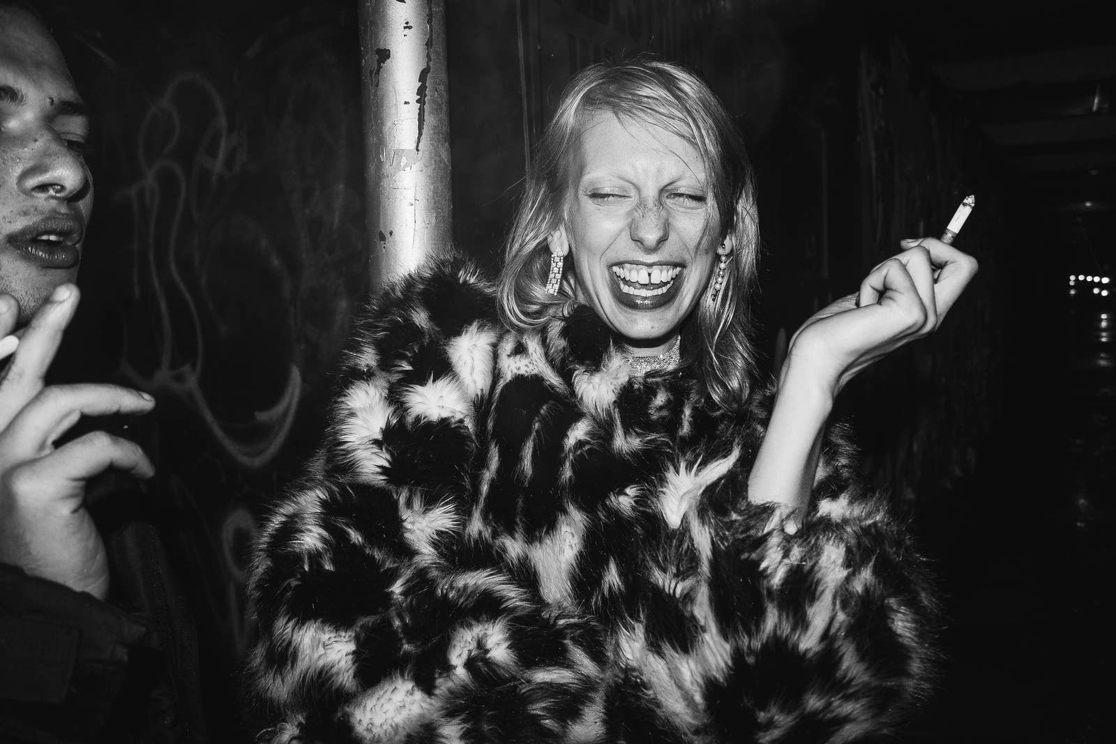 A woman wearing a fur coat laughs while smoking a cigarette