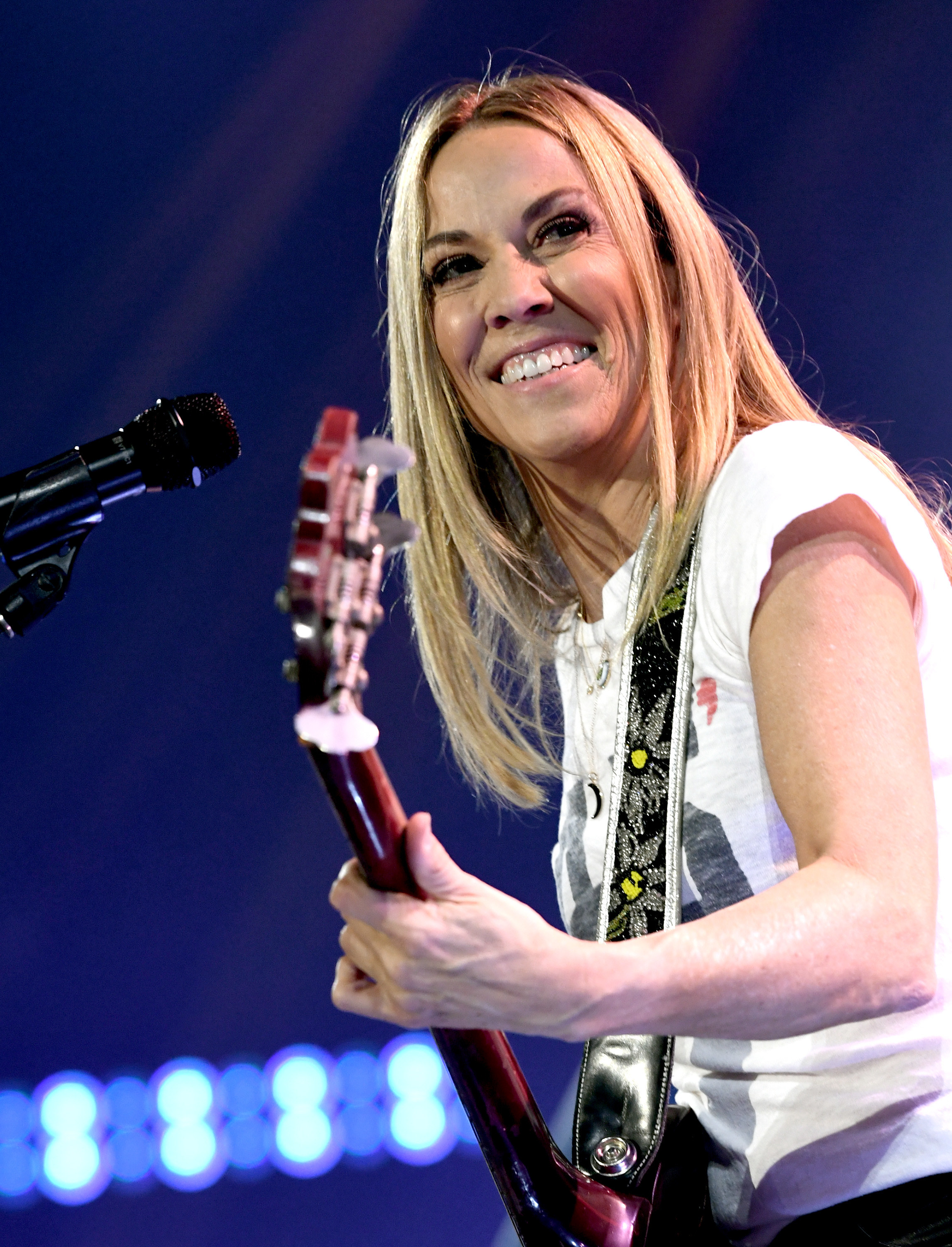 Sheryl Crow wearing a white t-shirt, holding a guitar and smiling.