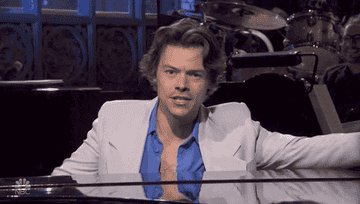 Harry Styles sitting at a piano on the set of SNL drinking from a martini glass