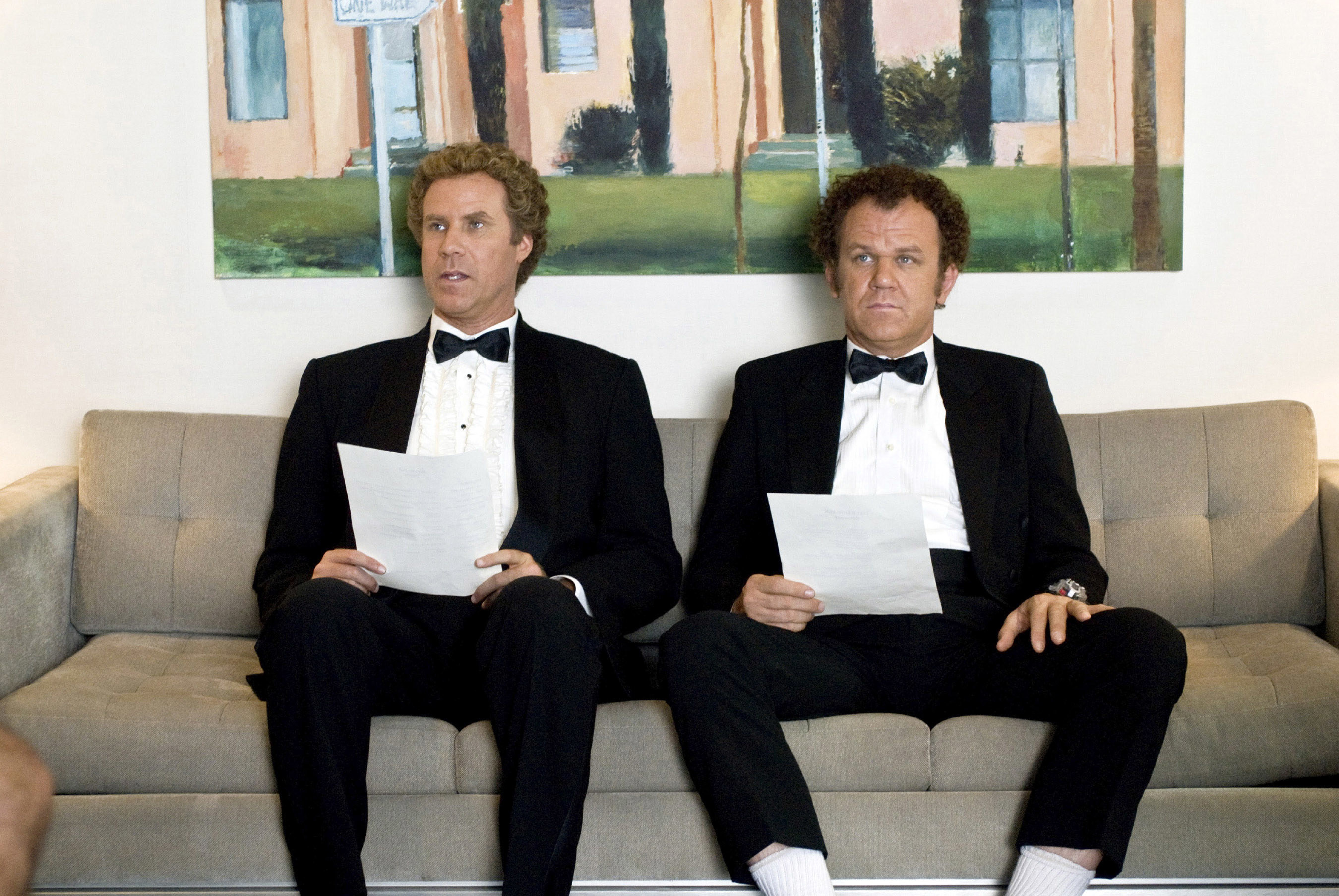Ferrell and Reilly hold resumes while sitting on a couch and wearing tuxedos