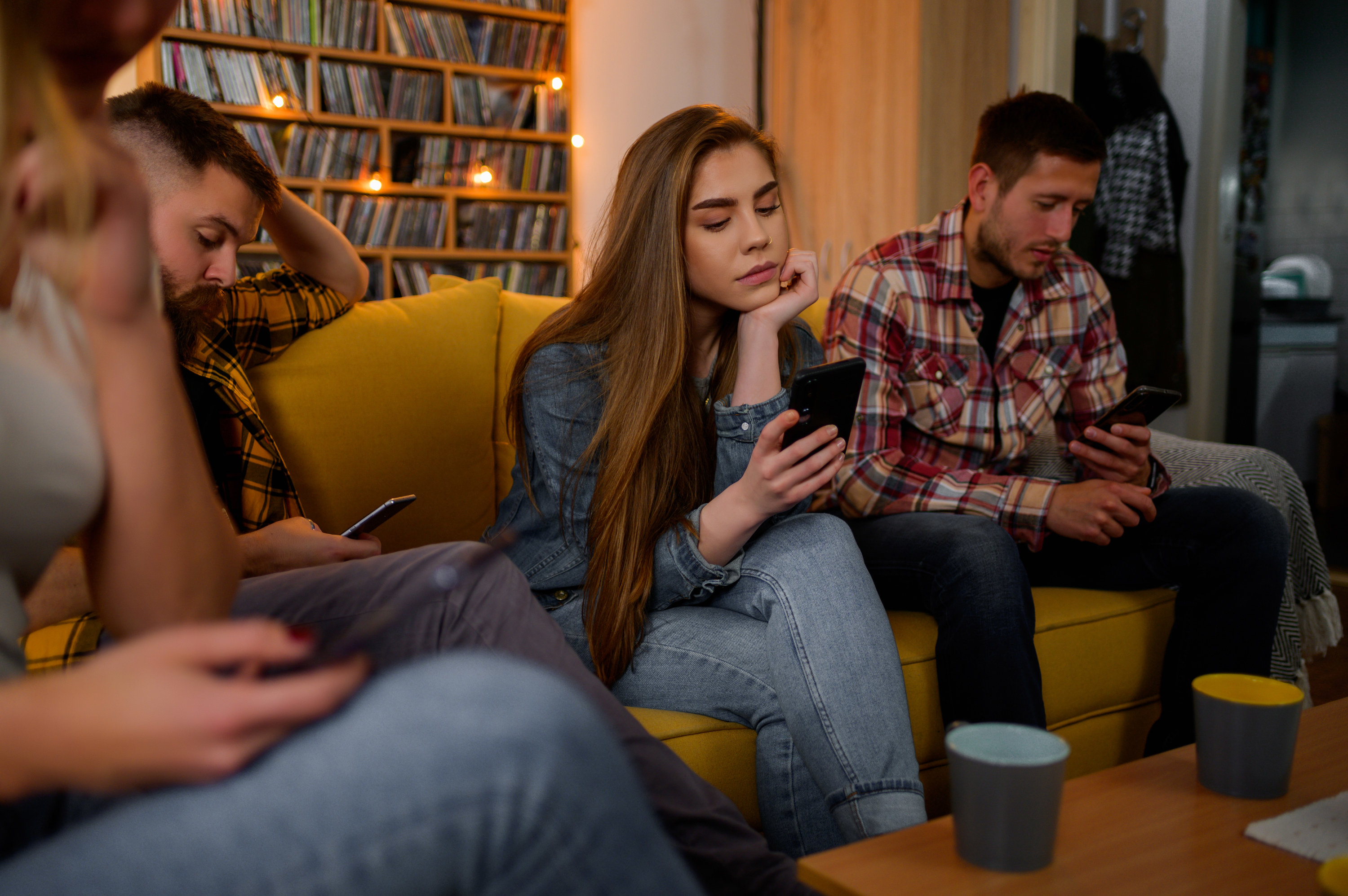A girl looking sad at her phone sitting on the couch surrounded by people