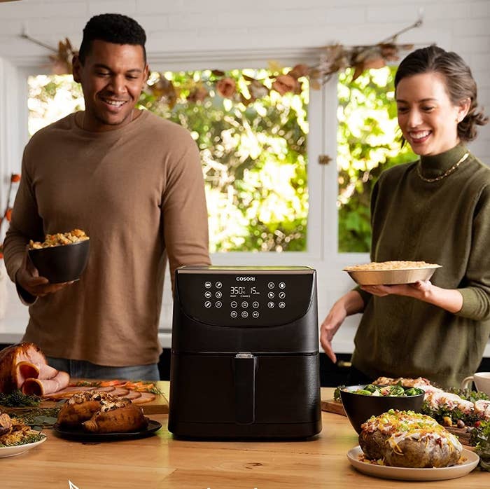 Two people on either side of the air fryer holding holiday goodies