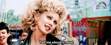 Blonde woman with red lipstick on, and a leather jacket saying &quot;Tell me about it, stud&quot;.