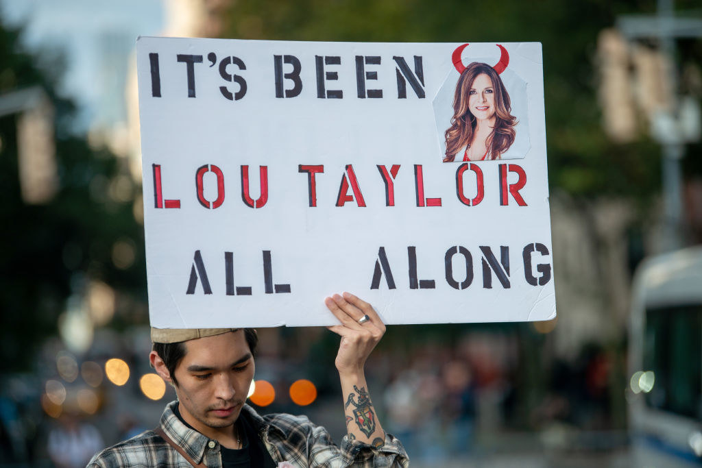 A person holding up a sign that says &quot;It&#x27;s been Lou Taylor all along&quot; with a photo of Lou with devil horns