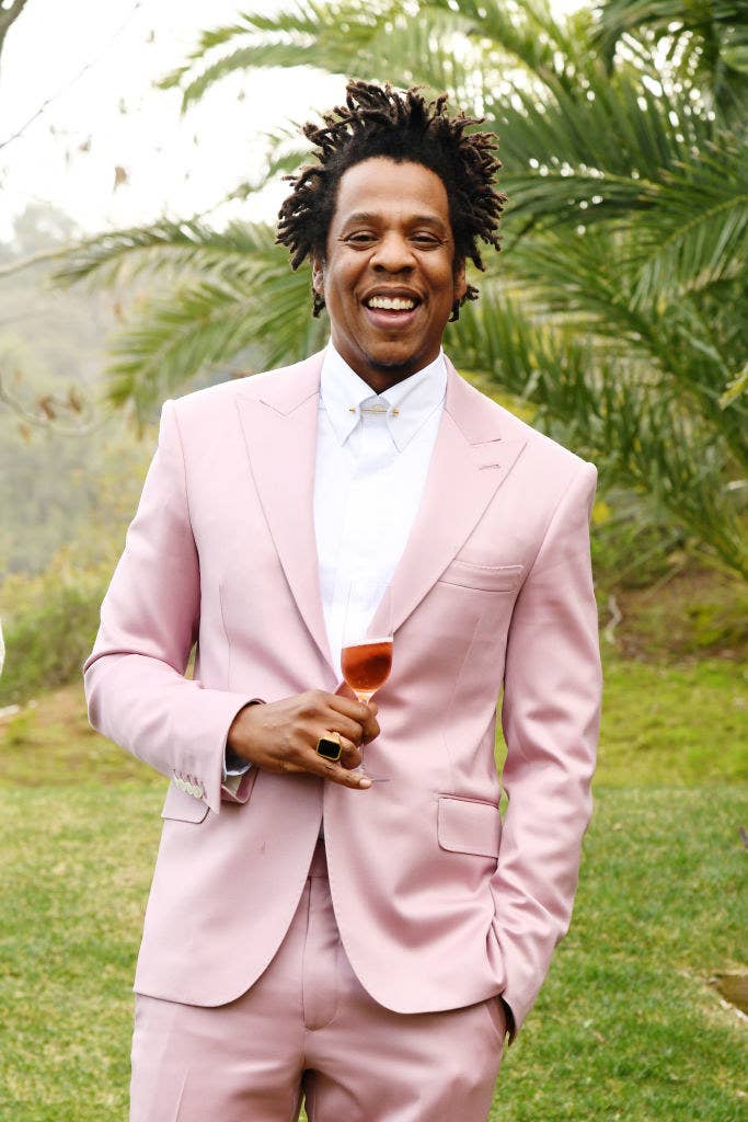 Jay-Z wearing a pastel suit and smiling while he stands outside with a glass of champagne