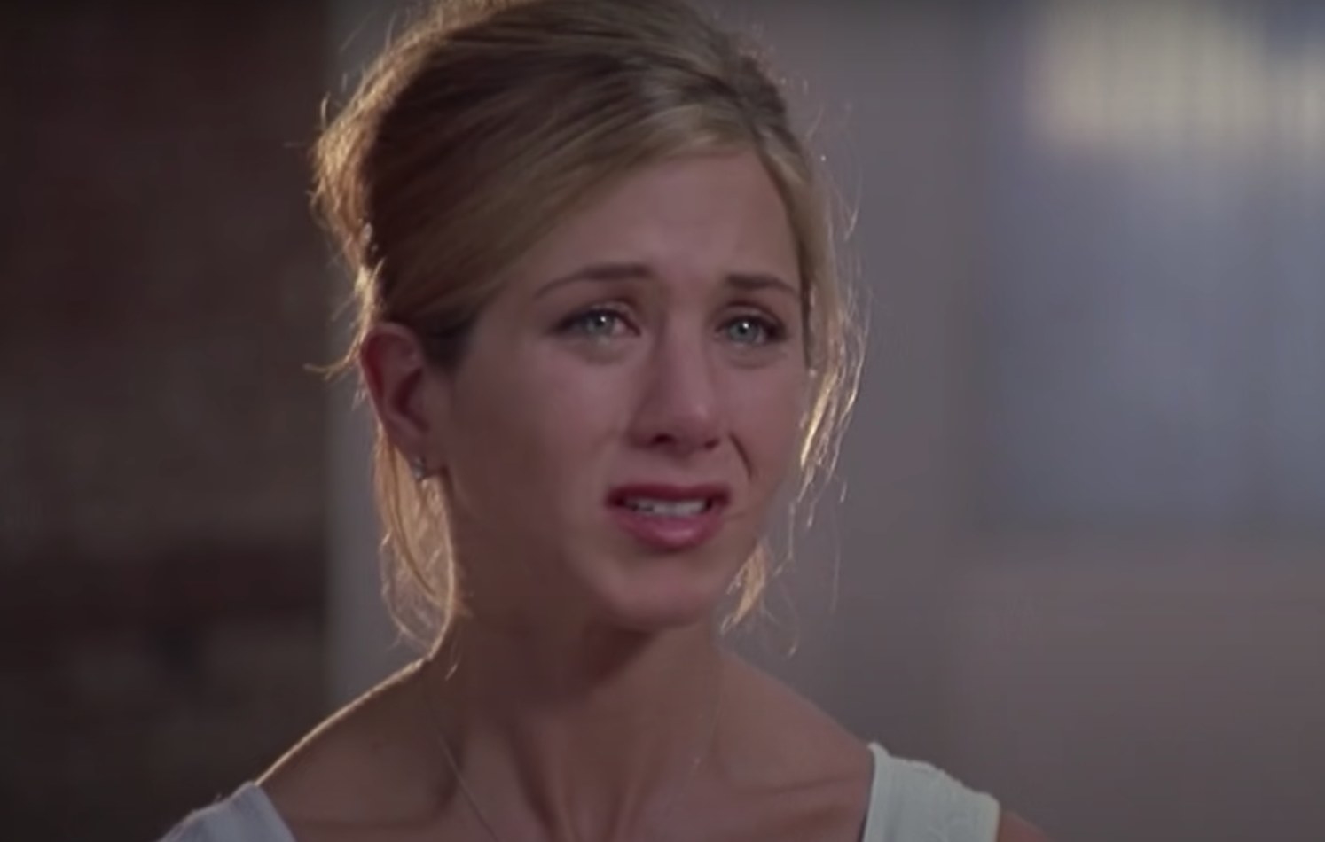 Jennifer Aniston has her hair in a bun and tears in her eyes while grimacing.