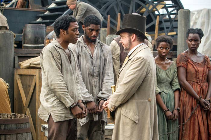 Chiwetel Ejiofor and Paul Giamatti speak on a dock surrounded by slaves