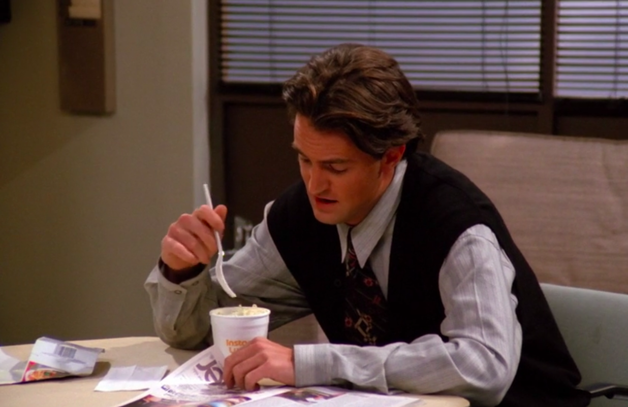 Chandler swearing pants, a long-sleeved shirt, a tie, and a sweater-vest, sitting at a table and eating something in a cup