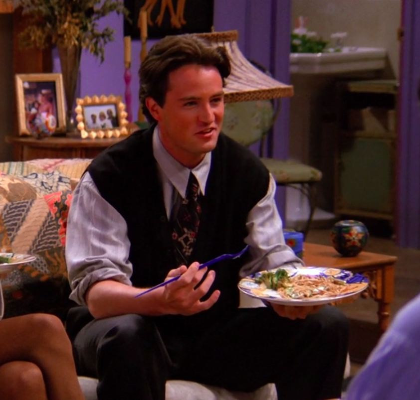 Chandler wearing pants, a long-sleeved shirt, a tie, and a sweater-vest, sitting on a couch, and eating something on a plate