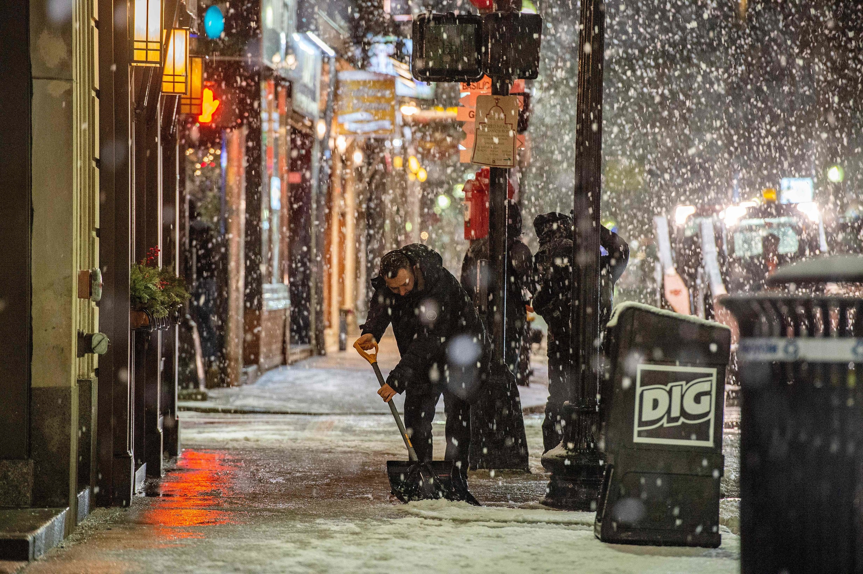 Snow falling outside a busy street or businesses, with someone shoveling the fallen snow