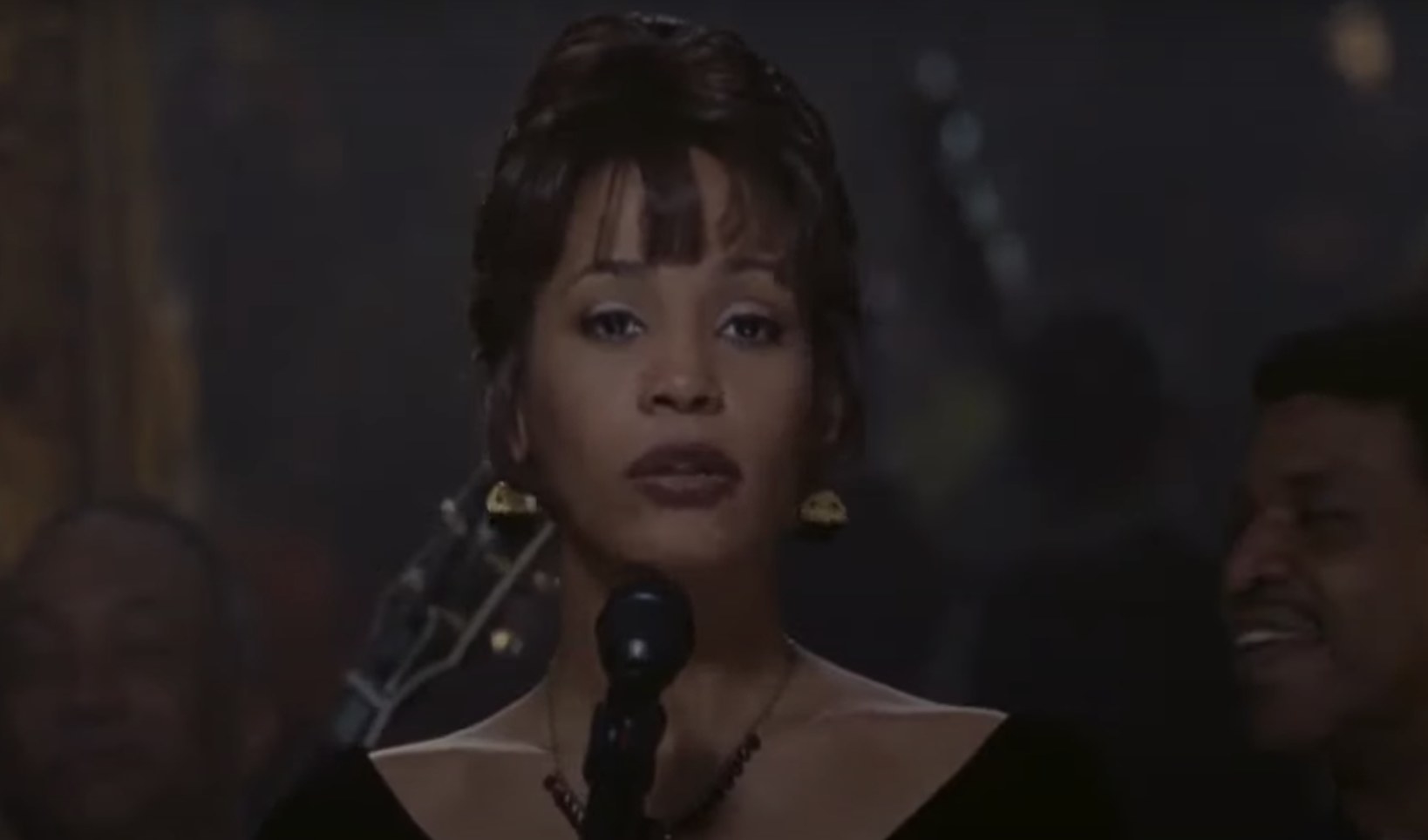 Whitney Houston stands on a stage with a microphone before her. She wears a black top and dangly earrings