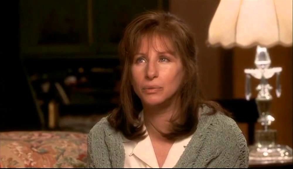 Barbra Streisand sits on a couch wearing a green water. She has a sad face and looks upwards offscreen as if she is upset.
