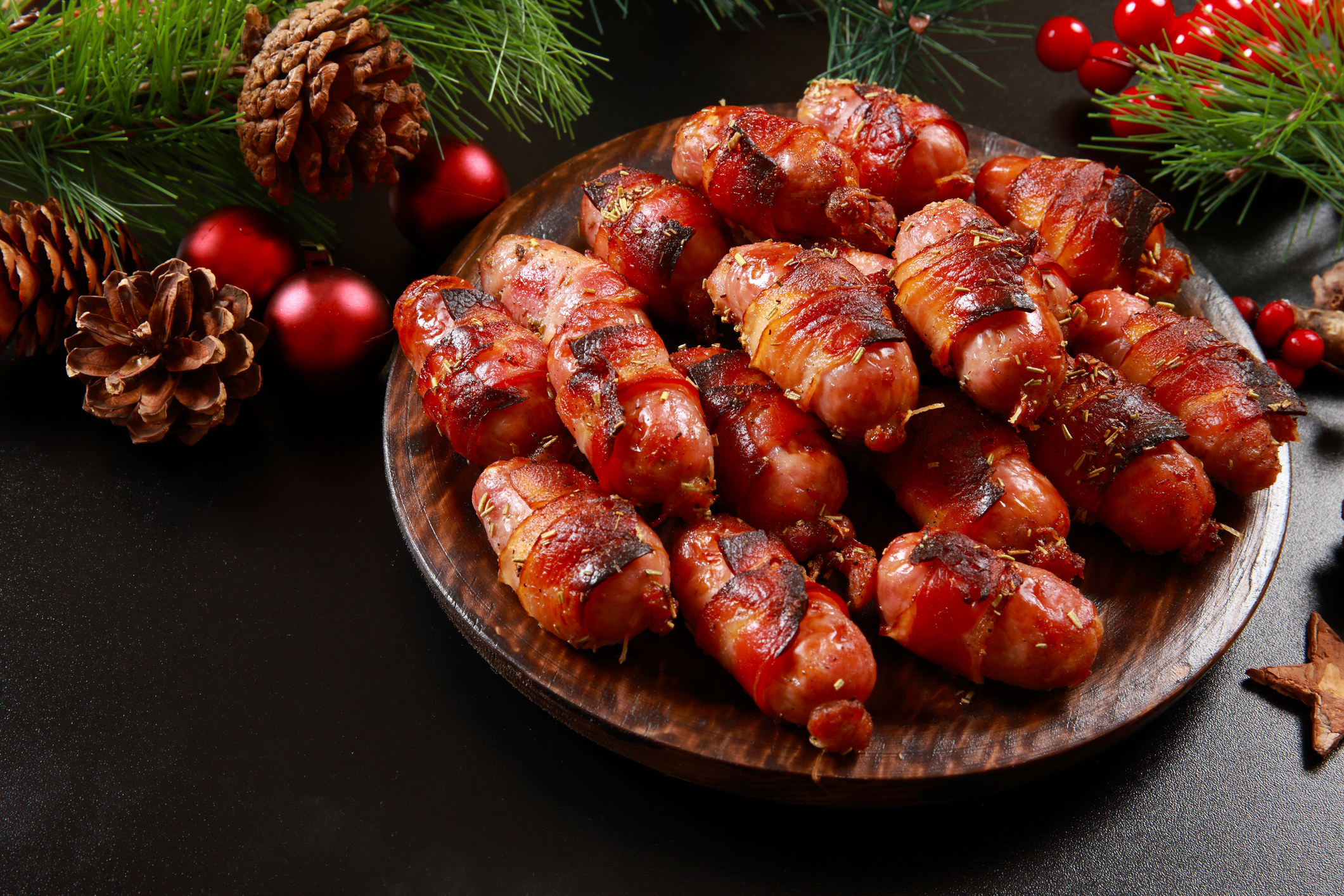 A plate of pigs in blankets (Sausages wrapped in bacon) with pine cones, holy berries and sprigs of tree around it.
