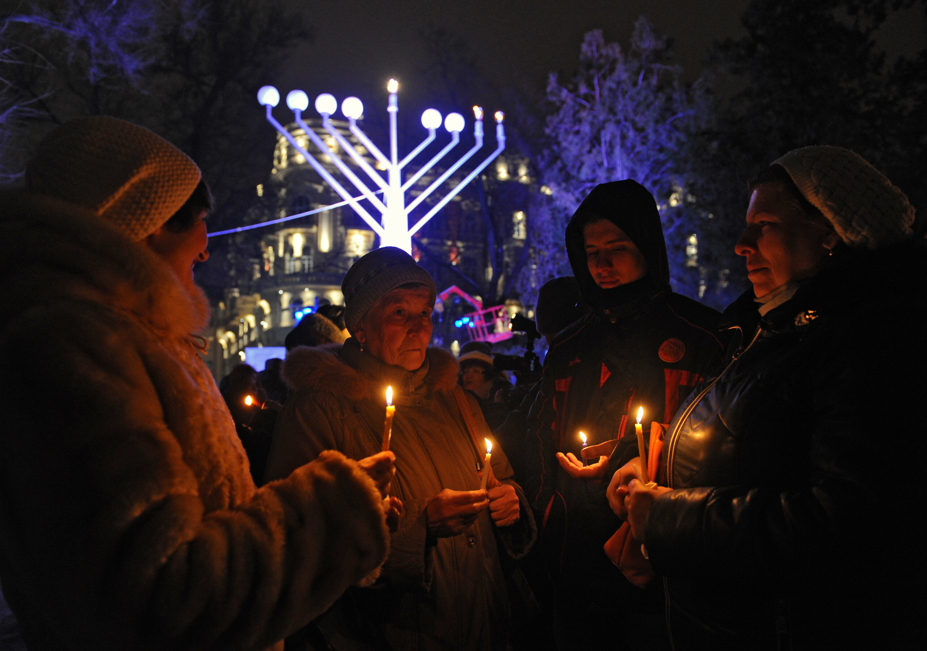 Four people hold candles in a semi circle outside at a Hanukkah celebration, a large menorah is behind them