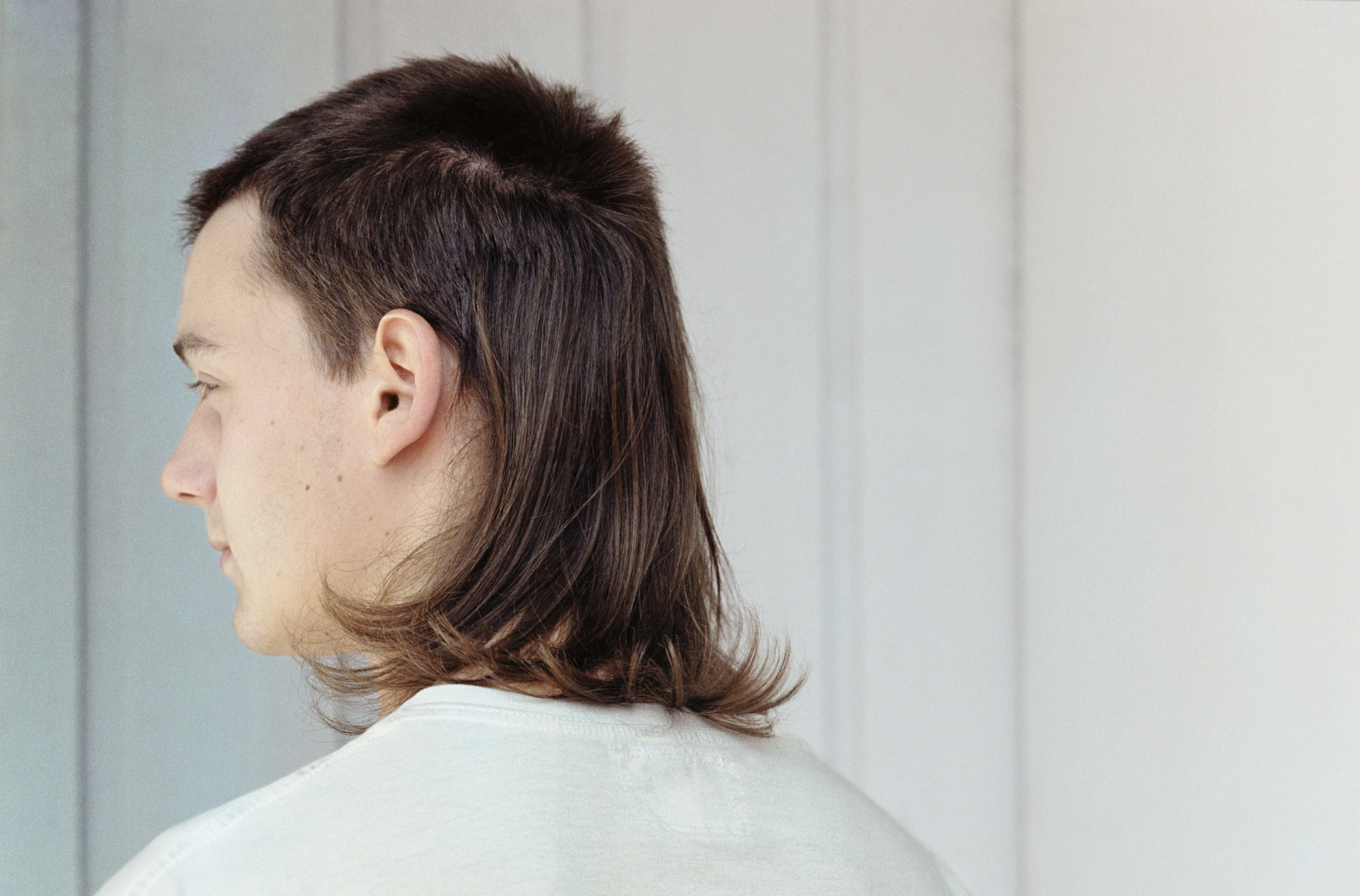 A photo of a man from behind who has a mullet and wearing a white tshirt.