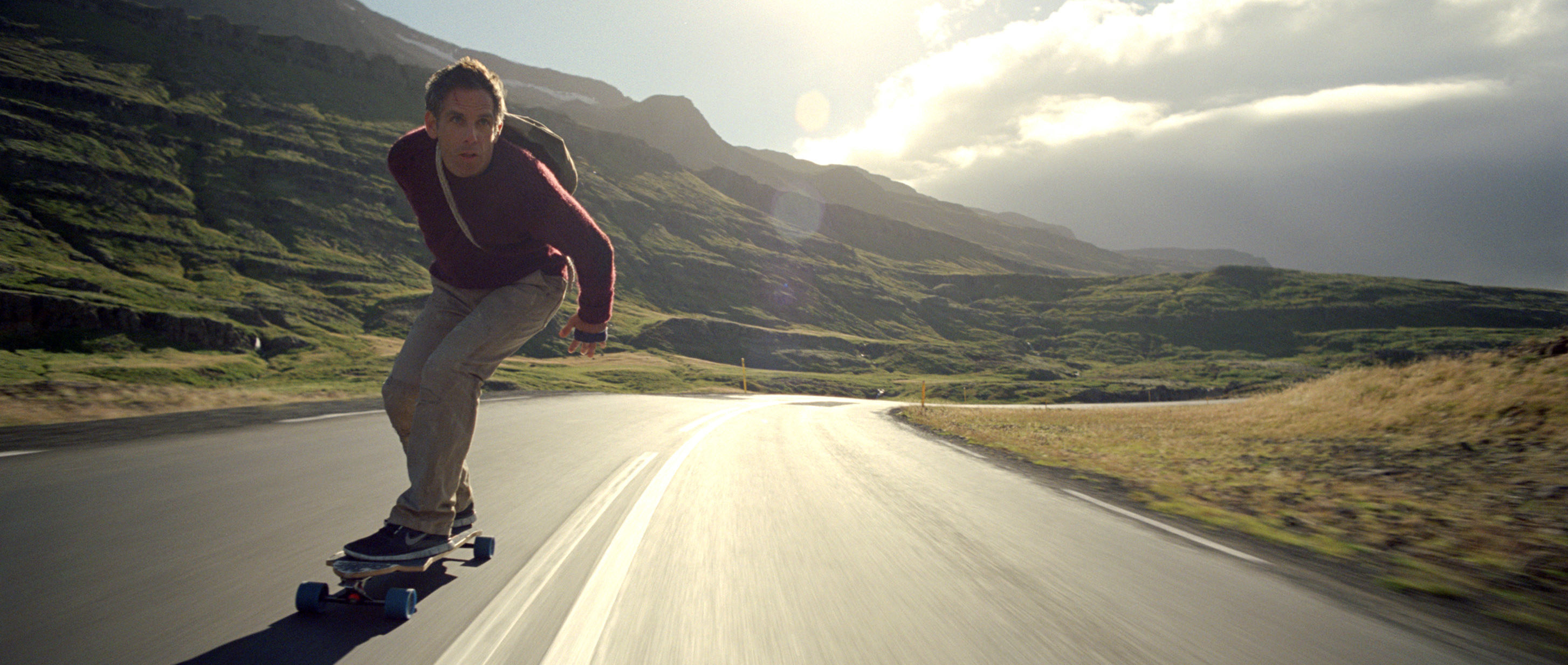 Ben Stiller riding a skateboard really fast down a quiet road in the countryside