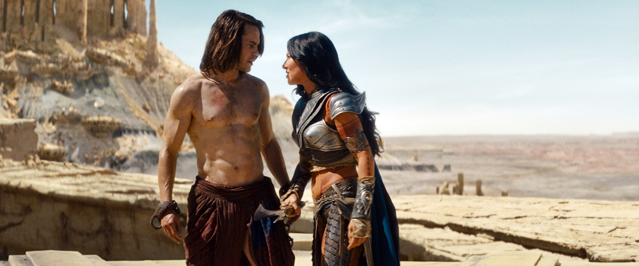 Taylor Kitsch, Lynn Collins standing on a desert planet out in space, holding hands