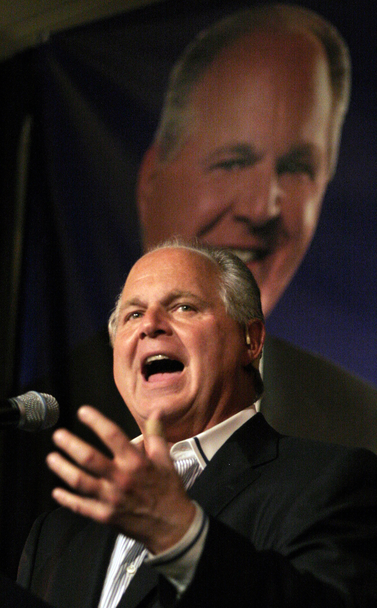 Rush Limbaugh in front of a photo of himself gesturing and with his mouth open in front of a microphone 