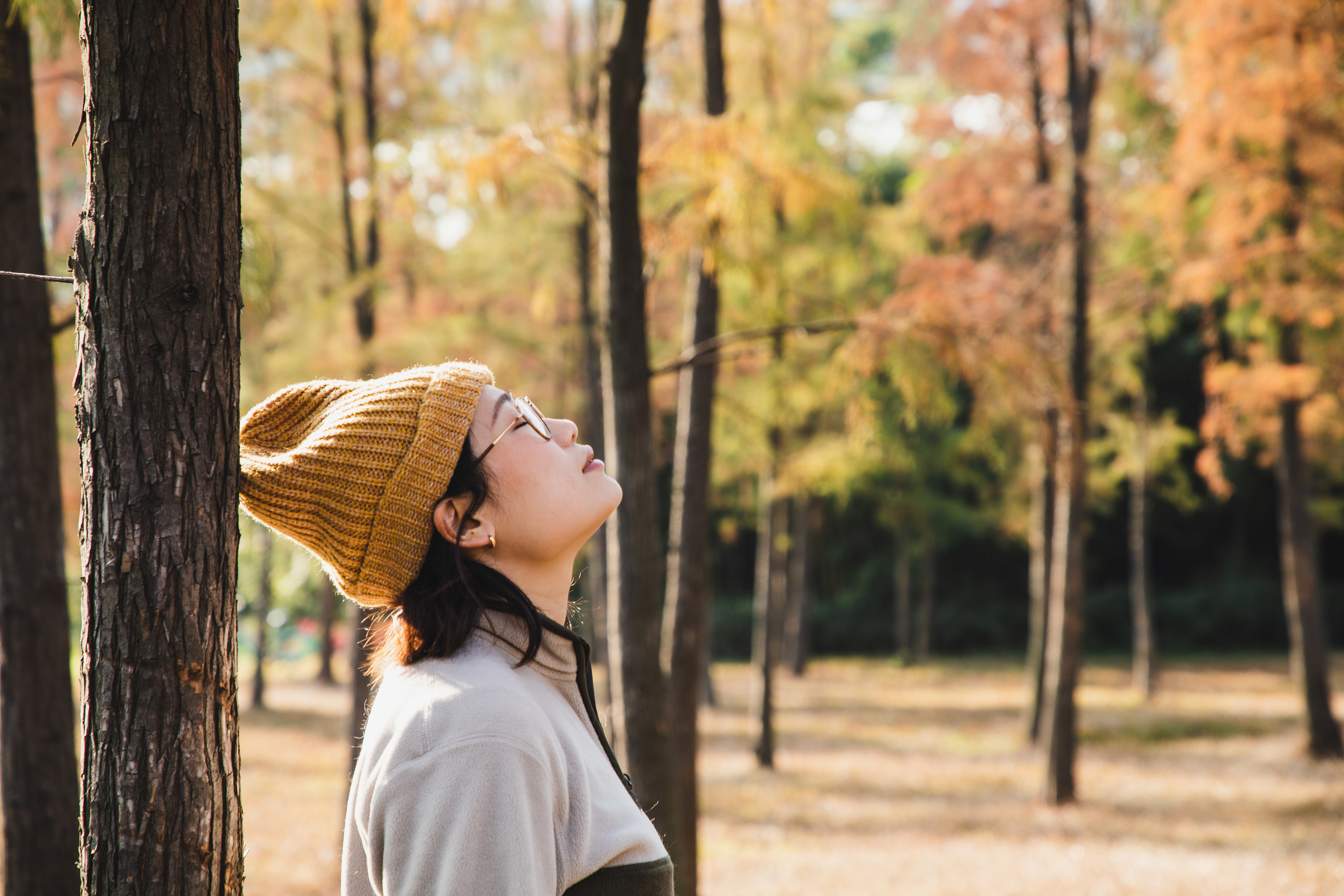 A person with a yellow beanie, glasses, earrings, and a jacket breathing in the air surrounded by fall trees