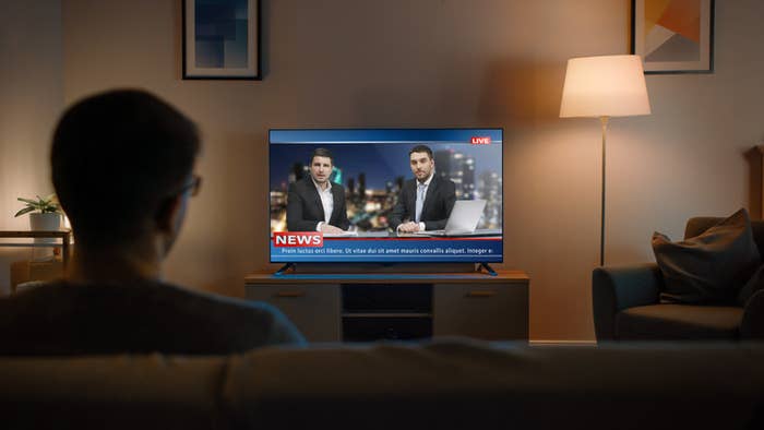 Young Man in Glasses is Sitting on a Sofa and Watching TV with Live News.