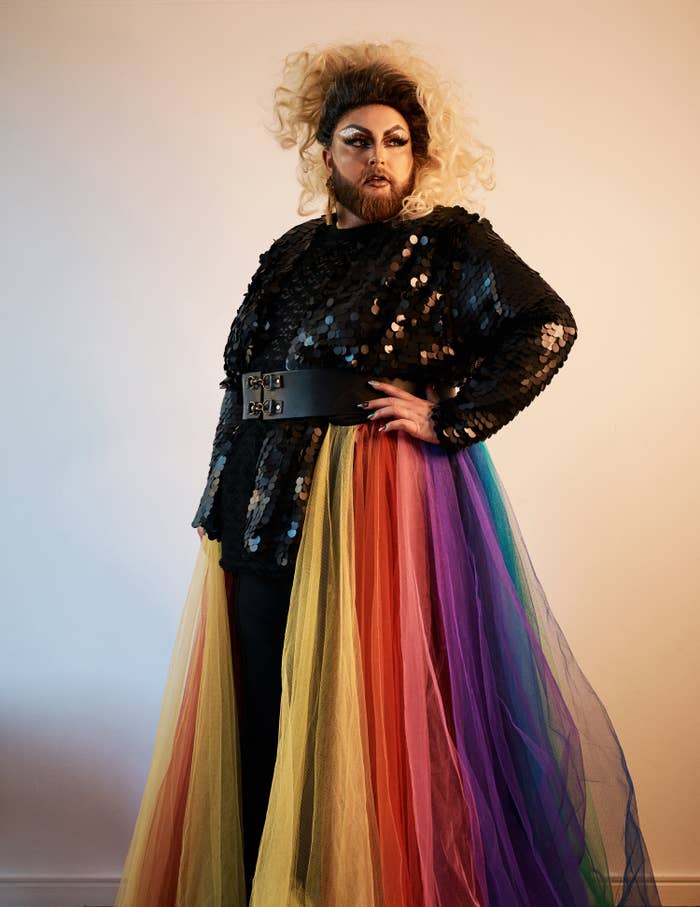 A stunning person in full makeup with a black sparkly top, a black belt, and a rainbow skirt. They have a brown beard and curly hair that changes from brown to blonde