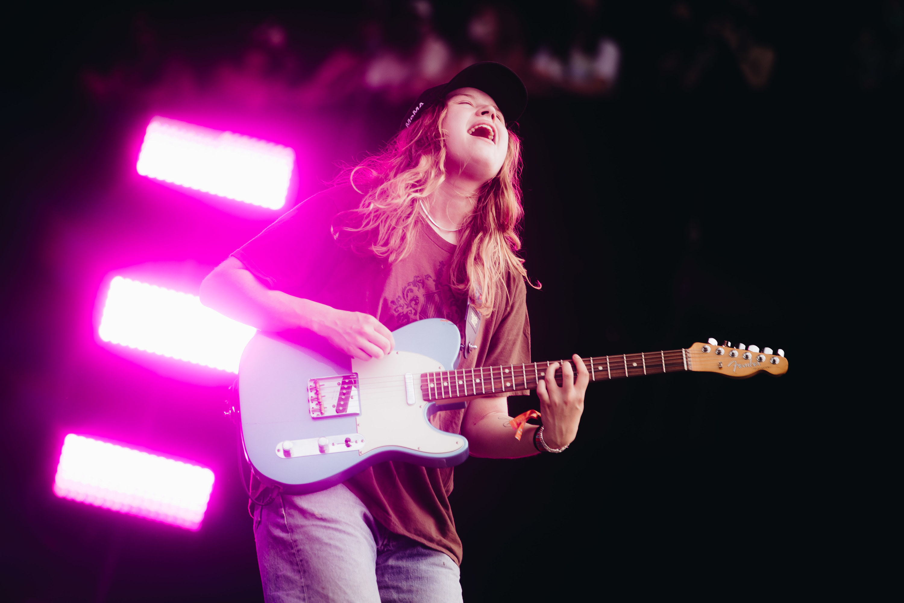 &quot;Girl In Red&quot; plying the electric guitar in singing with a brown t- shirt, blue jeans, and a black baseball cap. There is a blinding pink stage light behind her.