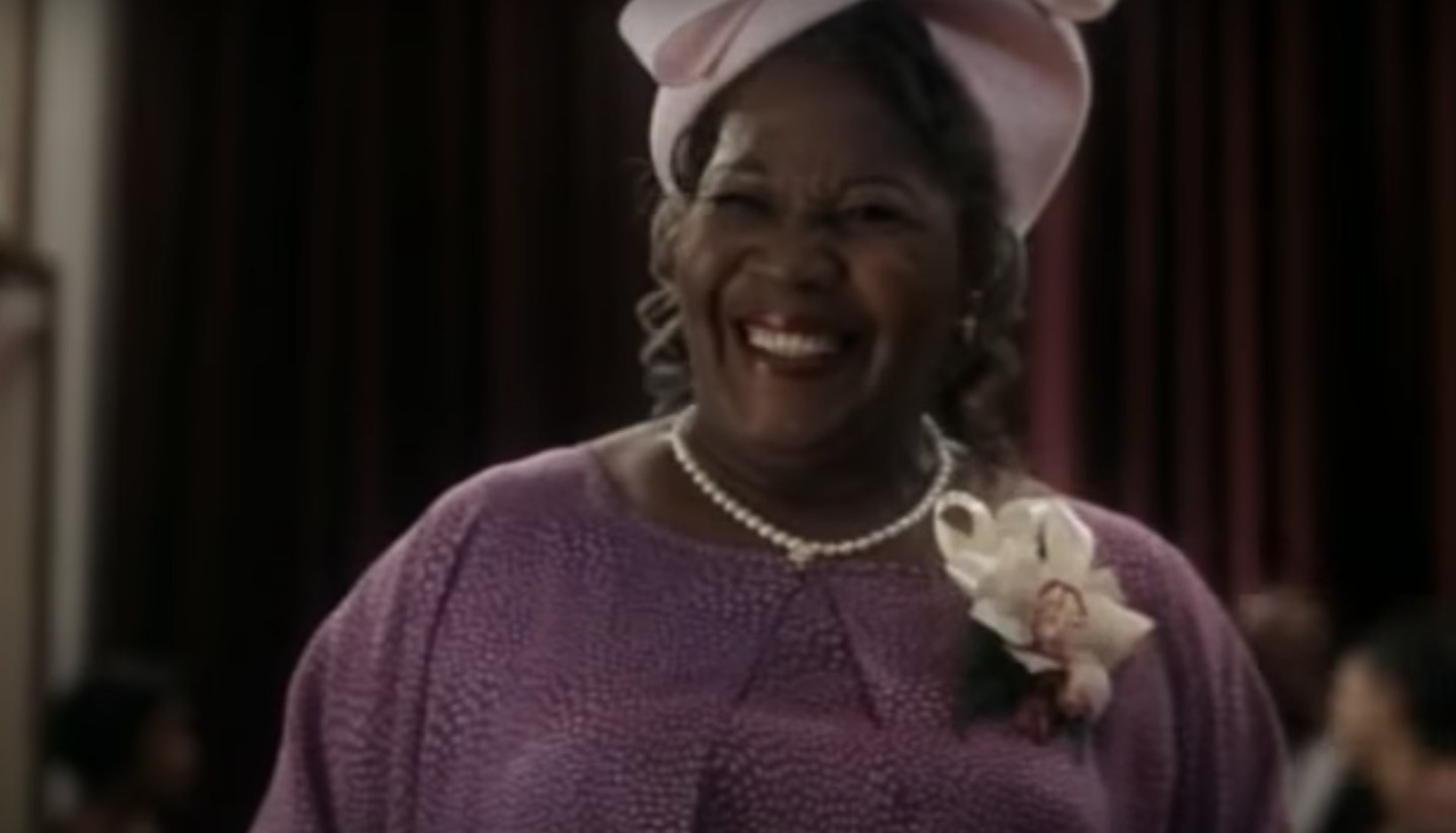 Actress Irma P. Hall wears a purple dress with pearls and a hat. She has on a very big smile