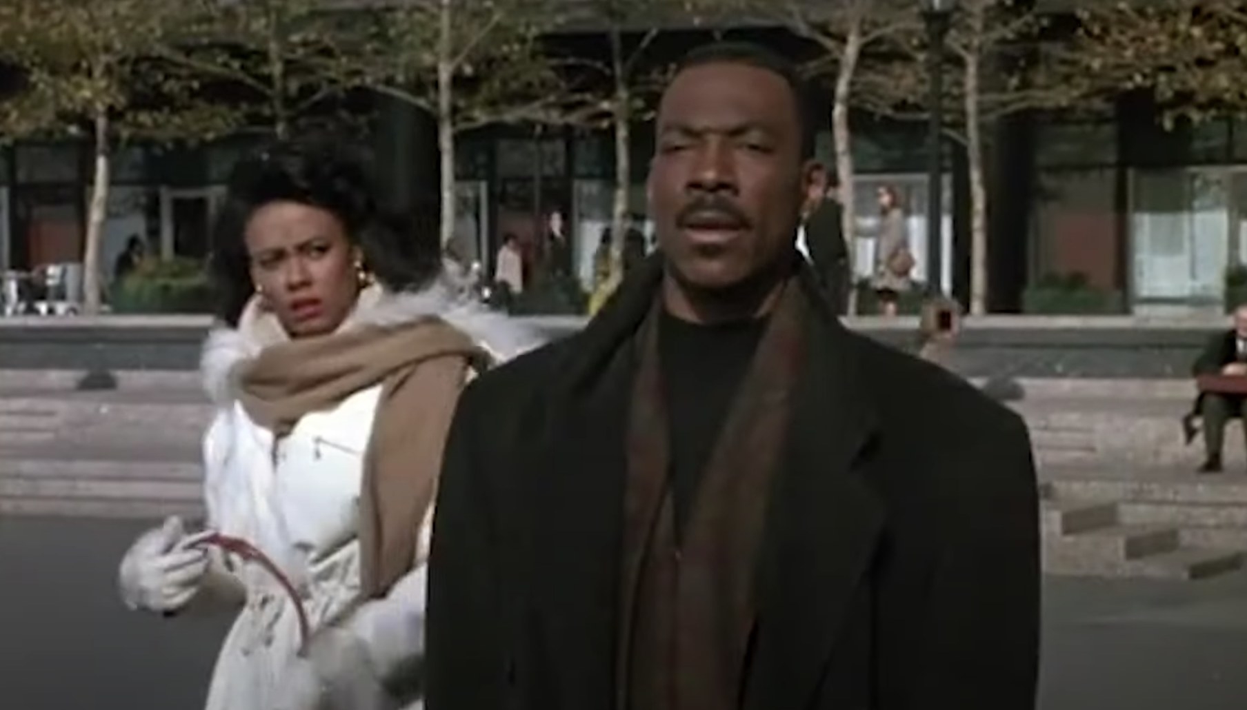 Actress Robin Givens stands behind actor Eddie Murphy while looking very confused. They are both wearing coats and are walking outside.