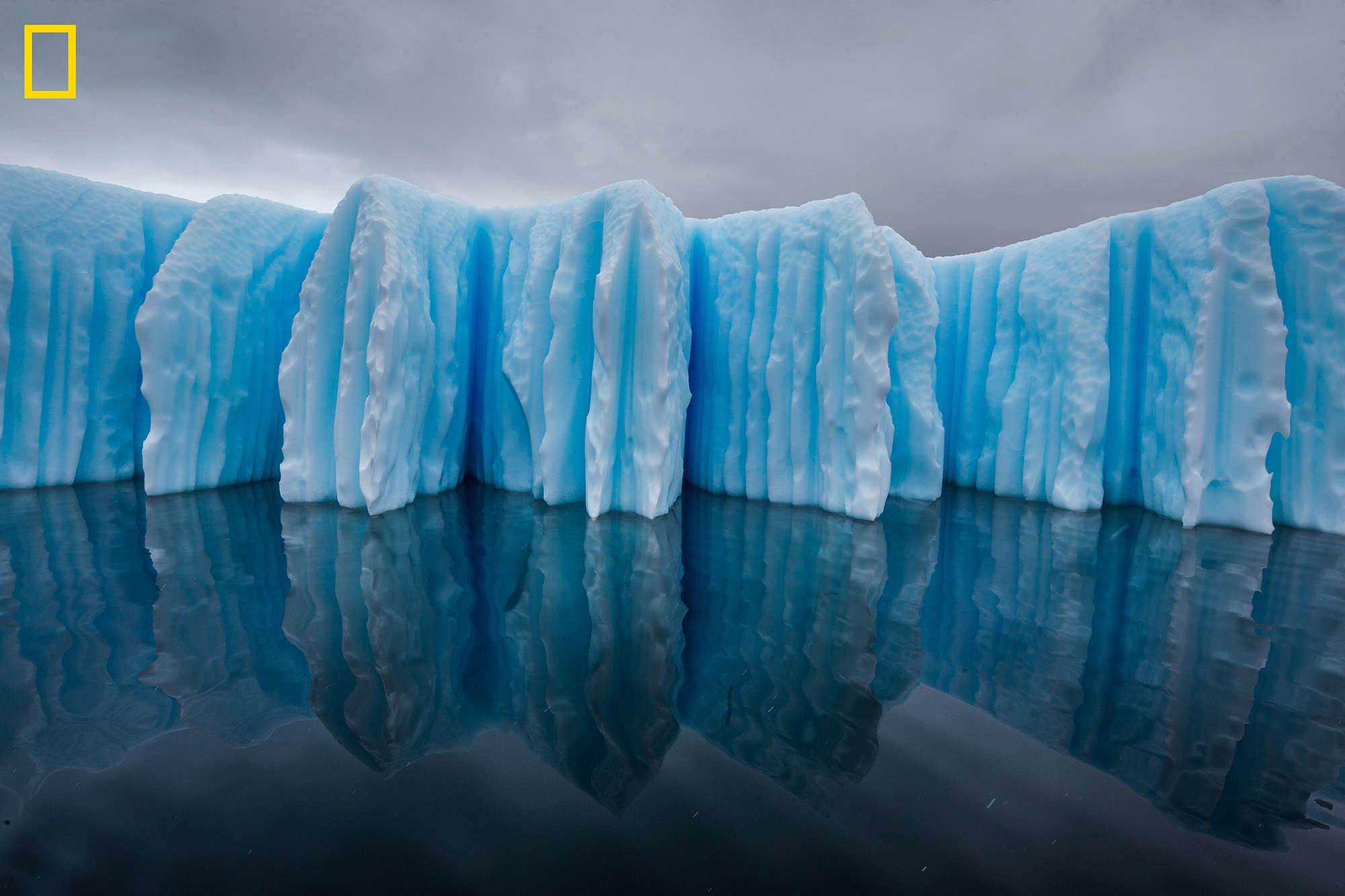 A massive glacier with vertical lines sits over the water under an overcast sky